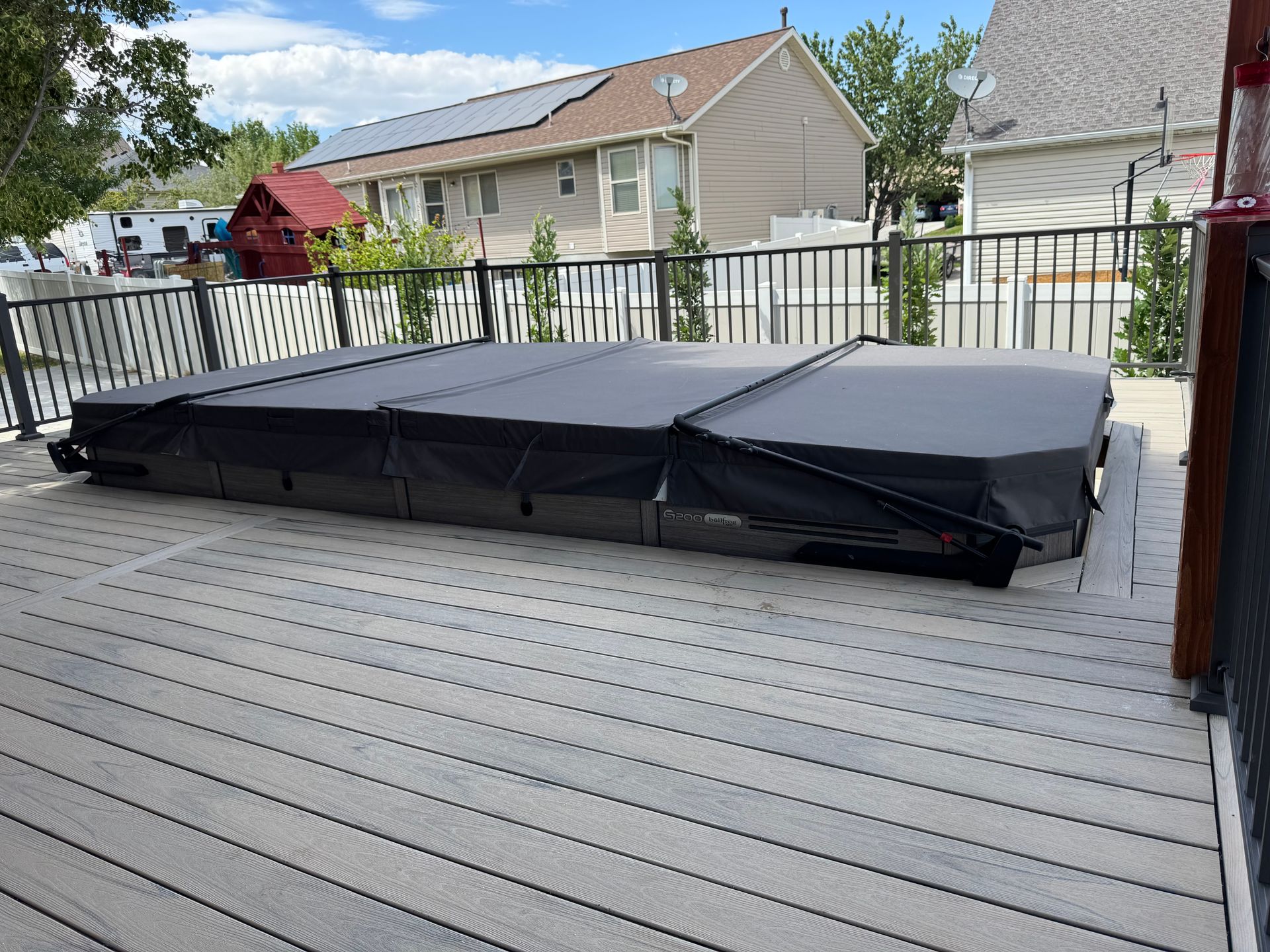 Black hot tub cover on a gray deck, surrounded by a black metal fence. Sunny day.