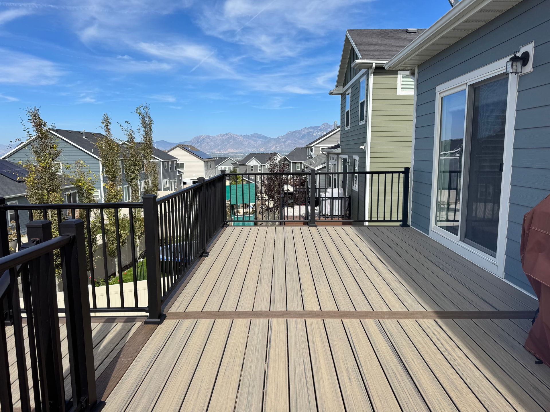 Wooden deck with black railing, overlooking a neighborhood and mountains under a blue sky.