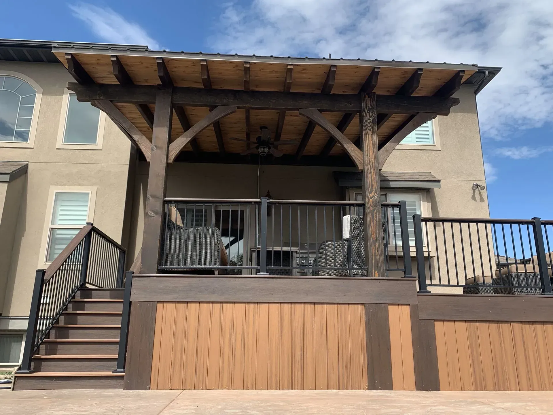 Wooden pergola over a composite deck with seating, built onto a light-colored house, under a blue sky.