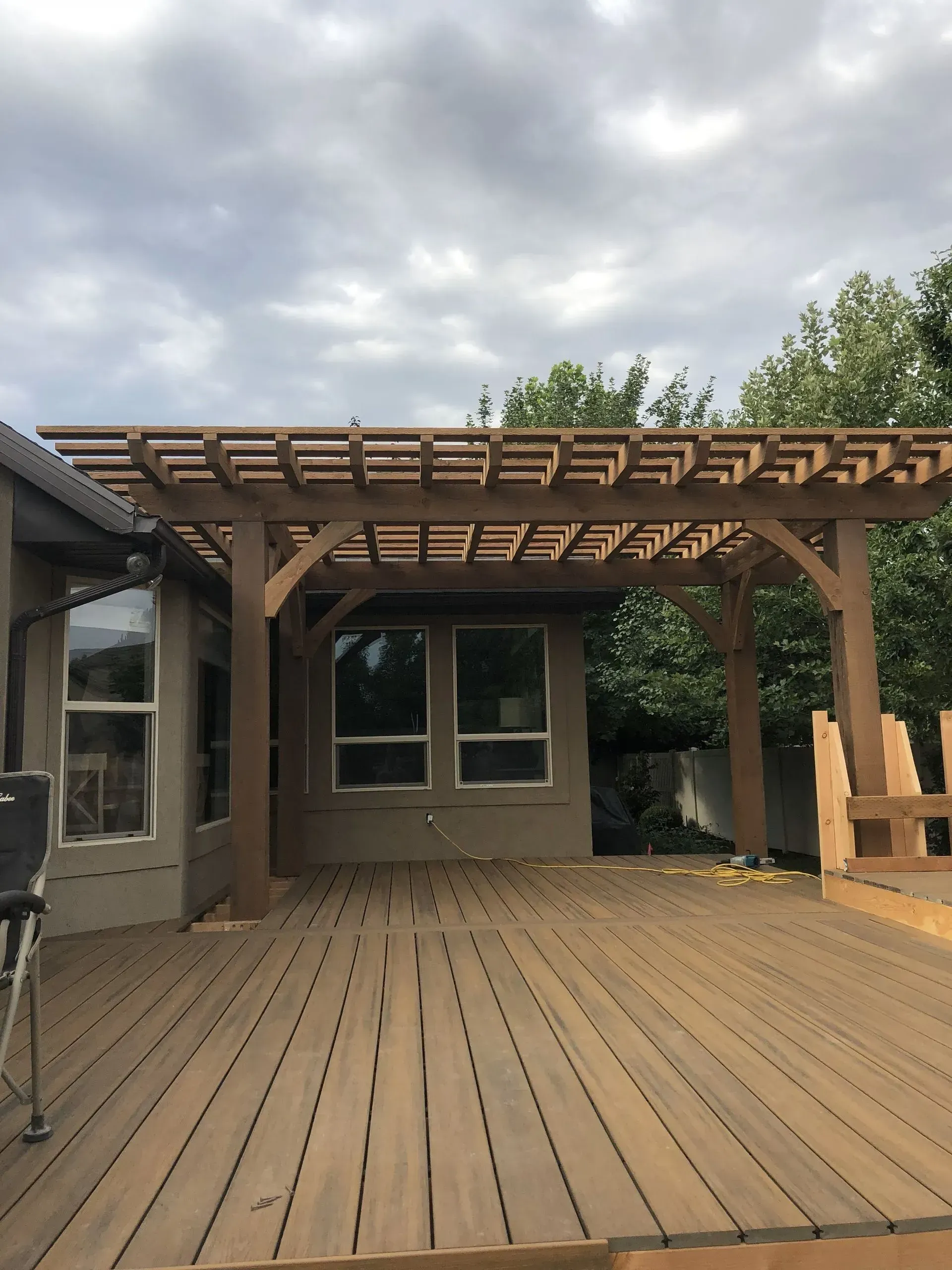 Wooden pergola over a deck extending from a house; brown wood, gray sky.