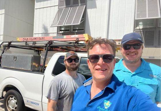 Three men in sunglasses pose in front of a white truck with tools and a ladder. The truck is parked near a white building with shutters.