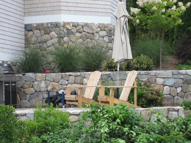 Two wooden chairs and an umbrella are in front of a stone wall
