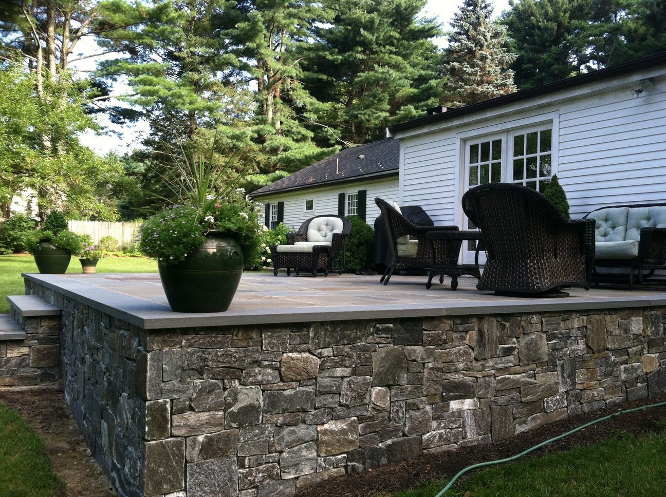 A stone wall surrounds a patio with a white house in the background