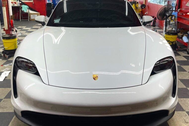 White Porsche sports car front view in a garage or service bay, with headlights visible and hood emblem centered