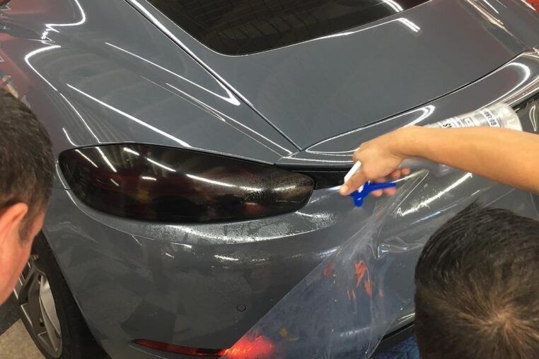 Hands applying a black vinyl wrap to a gray car panel with a squeegee