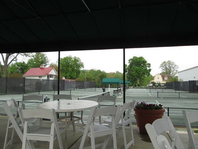 Outdoor view of tennis courts and white chairs around a table under a shaded patio.