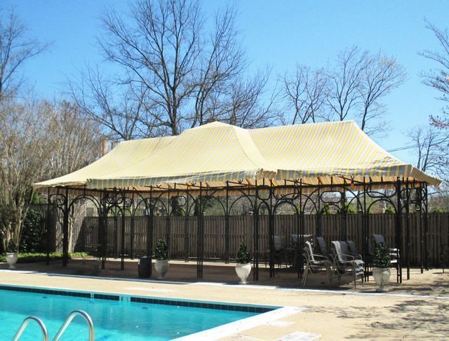Beige-roofed pergola by a swimming pool, with lounge chairs and trees in the background under a blue sky.