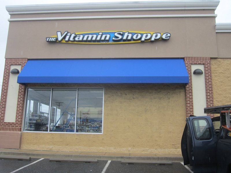 The Vitamin Shoppe store with blue awning, tan and brick exterior, and a truck parked outside.