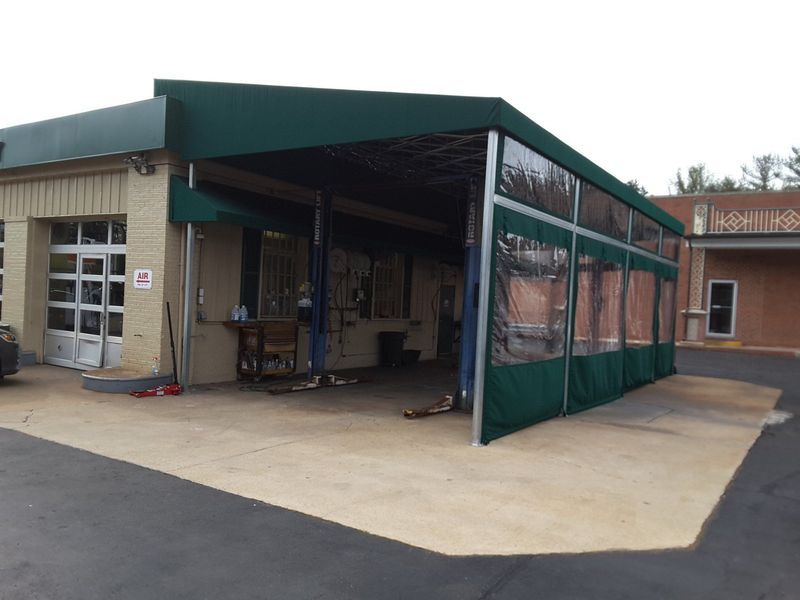 Green-roofed auto shop with roll-down side curtains. Lift, tools visible inside, beige brick exterior, concrete pad.