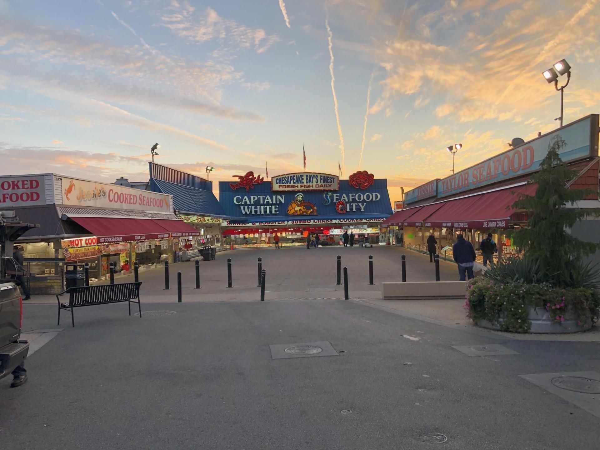 Outdoor seafood market with food stalls, blue and red awnings, under a sunset sky.