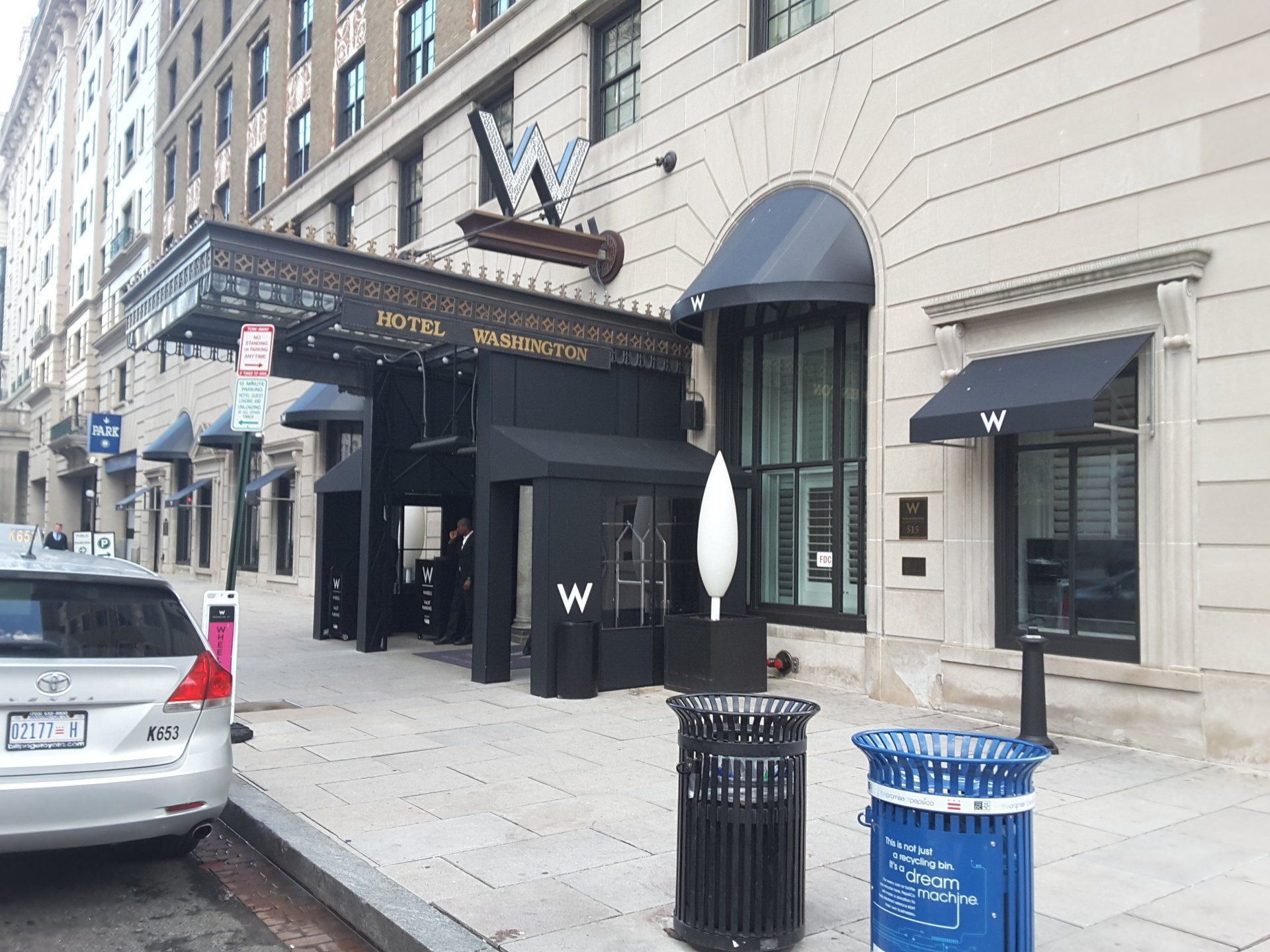 Hotel entrance with black awning, a white building, a street, and a car parked on the side.