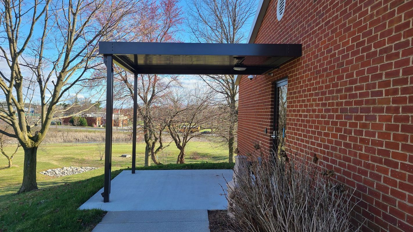 Black pergola over concrete patio attached to brick building; trees and yard in background.