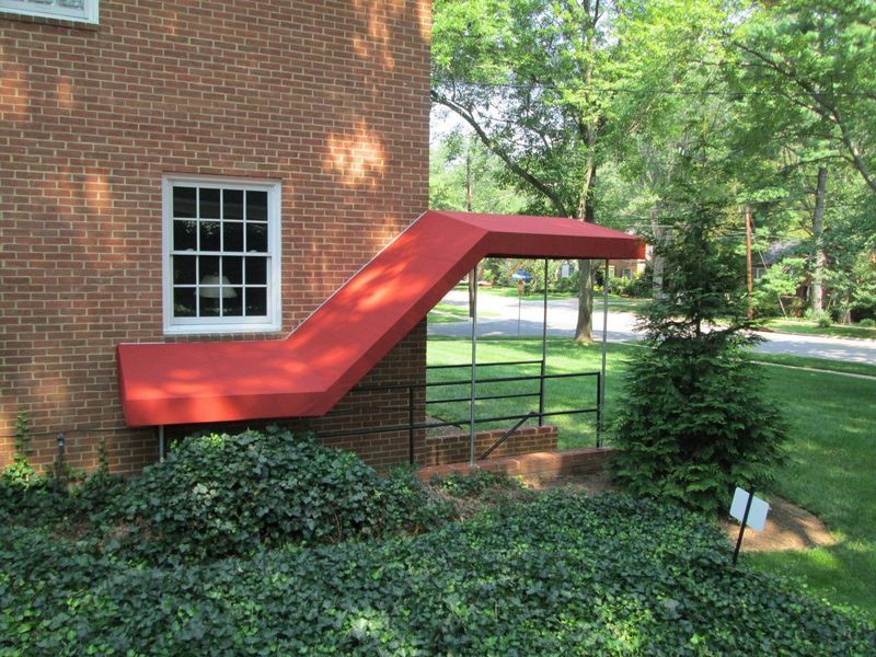 Red awning over entrance stairs of a brick building, with a window and greenery.