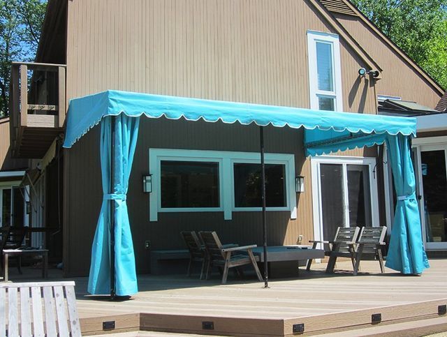 Teal canopy with curtains on a wooden deck, beside a brown house with large windows.