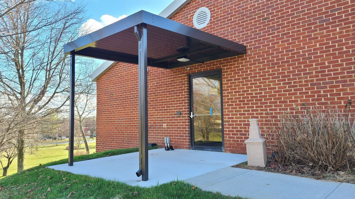 A metal canopy over a doorway on a brick building, supported by black posts, on a concrete patio.
