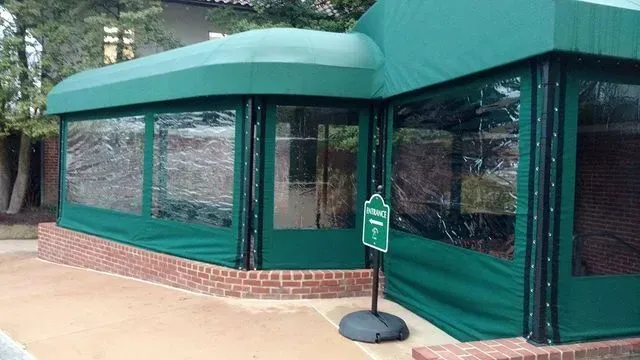 Green canopy-covered outdoor seating area with clear vinyl panels, brick base, and a sign.
