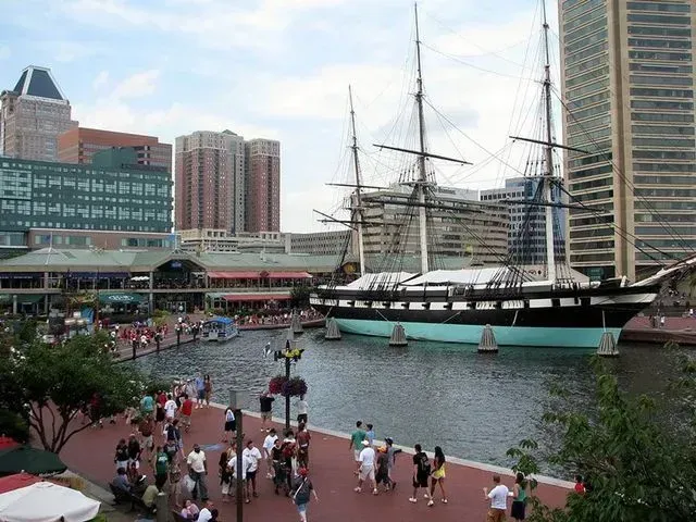 Baltimore Inner Harbor with tall ship, waterfront shops, and people walking. City buildings in the background.