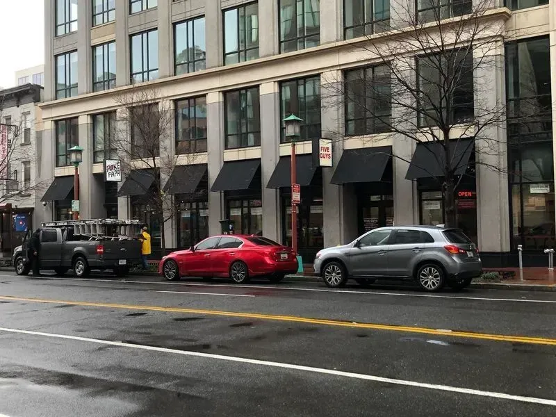 Cars parked on street in front of a building with awnings. A truck and a red sedan are visible.