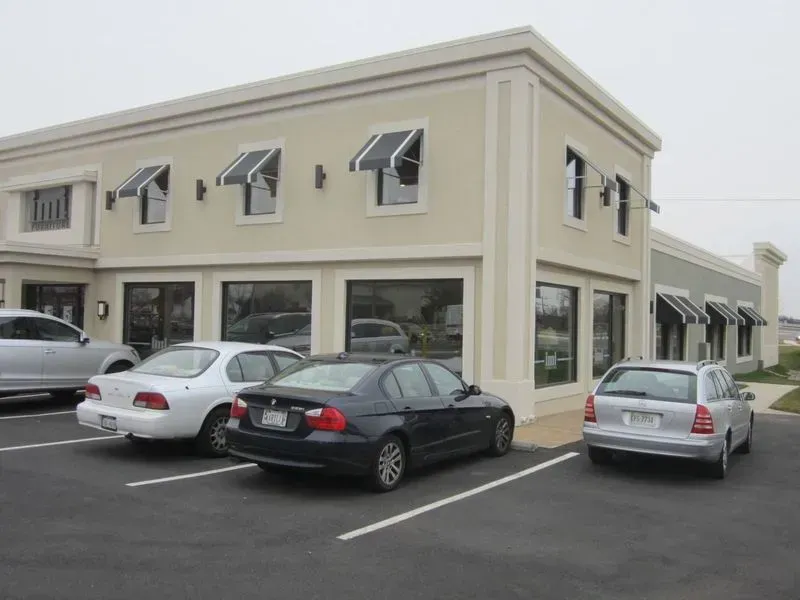 A two-story beige building with black awnings and large windows, cars parked in front.