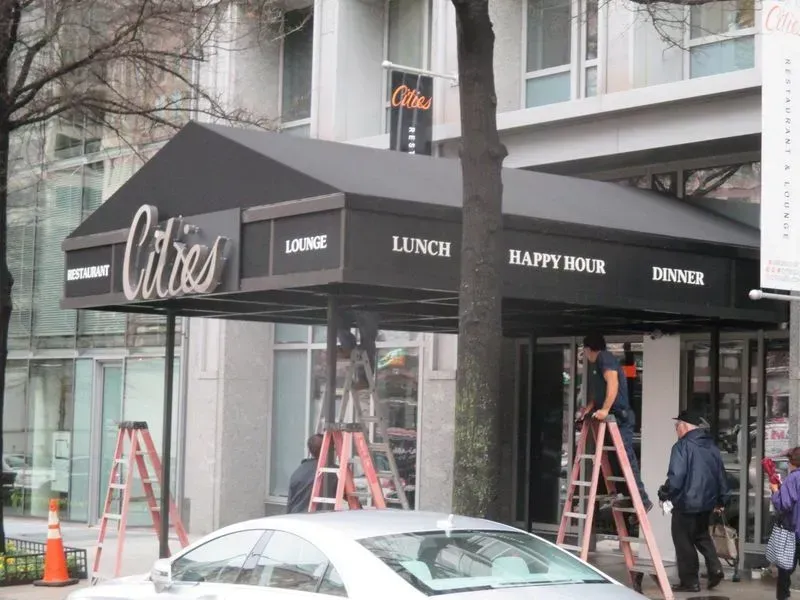 Exterior of Cities restaurant with workers, black awning, and a silver car.