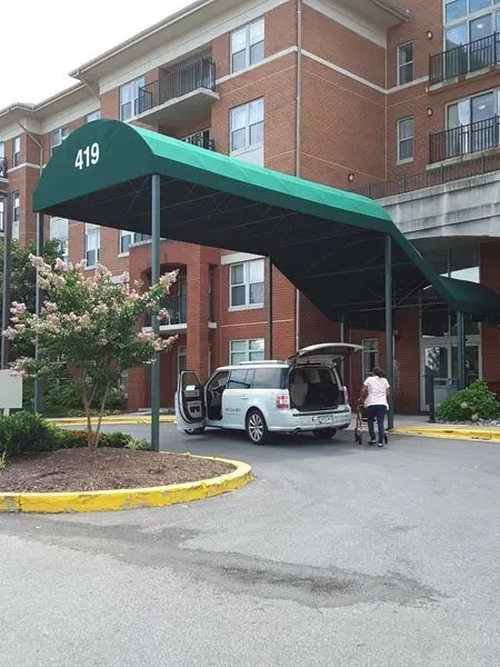 A silver van parked under a green canopy at an apartment building entrance, a person loads luggage.