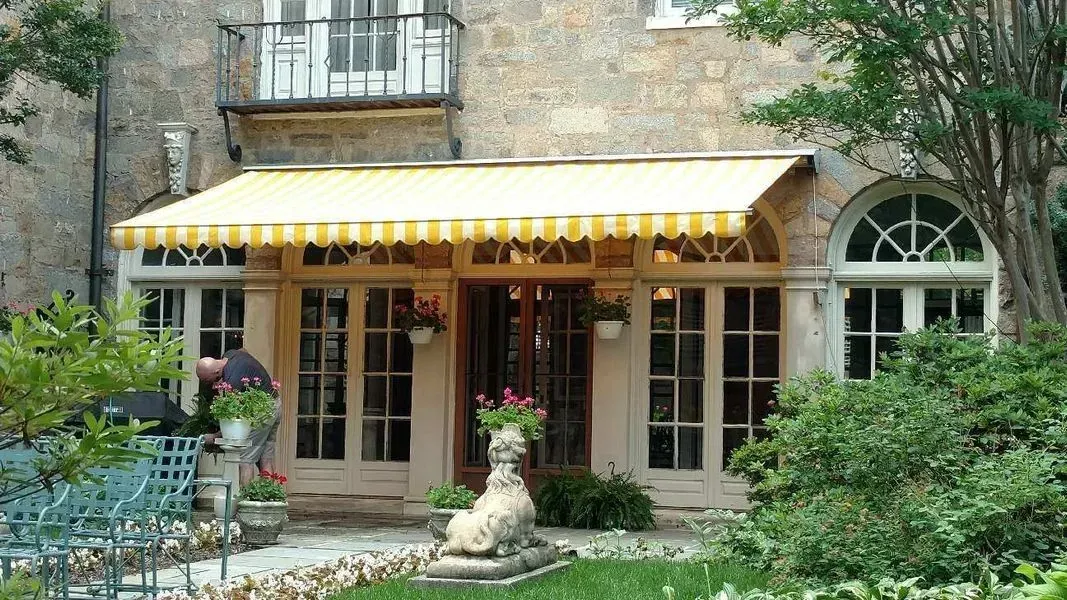 Exterior of a building with arched windows and a yellow striped awning. A stone statue and greenery are in front.