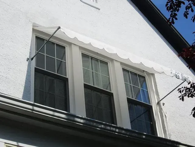 White building with three windows, each with a dark frame and white scalloped awning supported by metal arms.