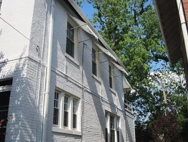 White two-story brick building with multiple windows. Overhead wires and green trees are visible.