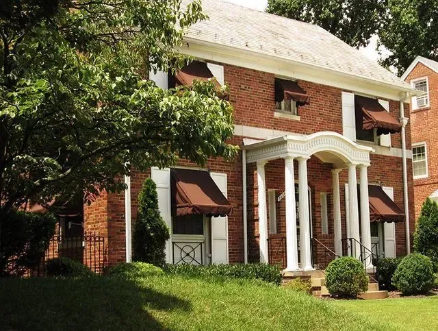 Two-story brick building with white columns and brown awnings. Green lawn and trees in front.
