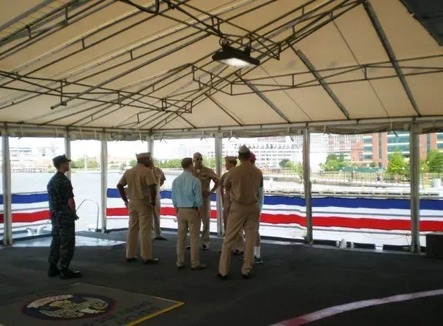 Navy officers gather under a tent on a ship deck with a waterfront view.