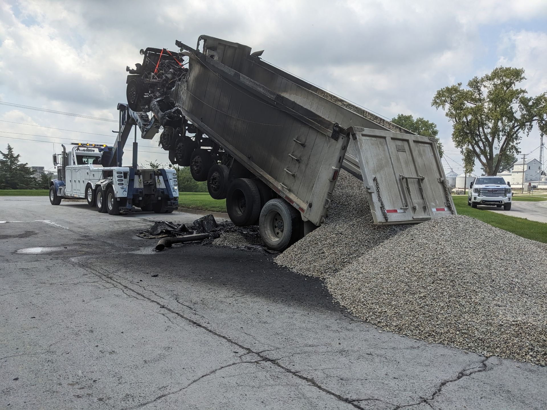 Dump truck overturned, spilling gravel onto a paved road; a tow truck attempts recovery.