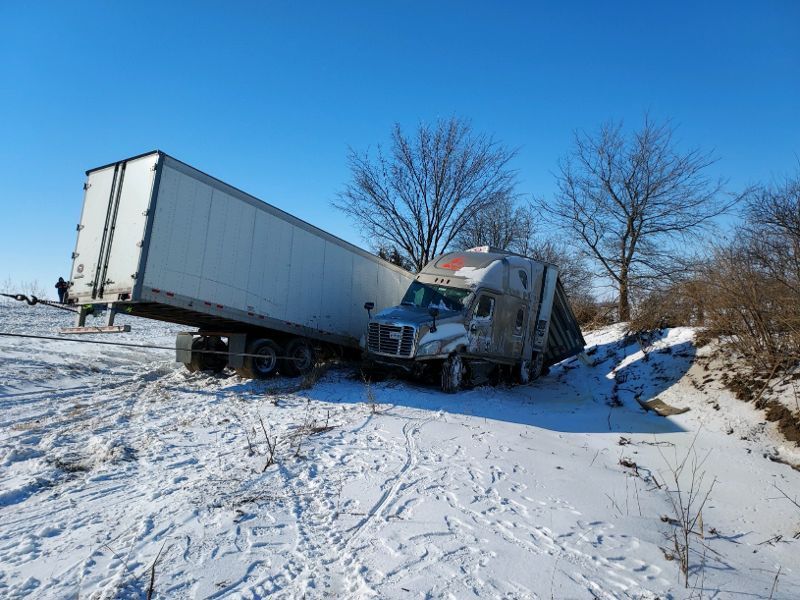 Semi-truck crashed off road, trailer leaning against the cab in a snowy ditch on a sunny day.