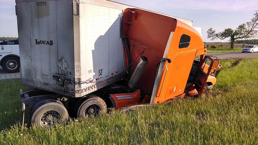 Semi-truck crashed off road, orange cab wedged against white trailer in grassy field.