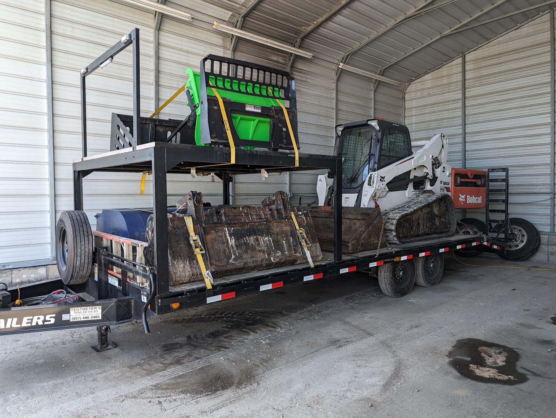 A trailer loaded with construction equipment inside a building. Includes a Bobcat, other tools, and a green attachment.