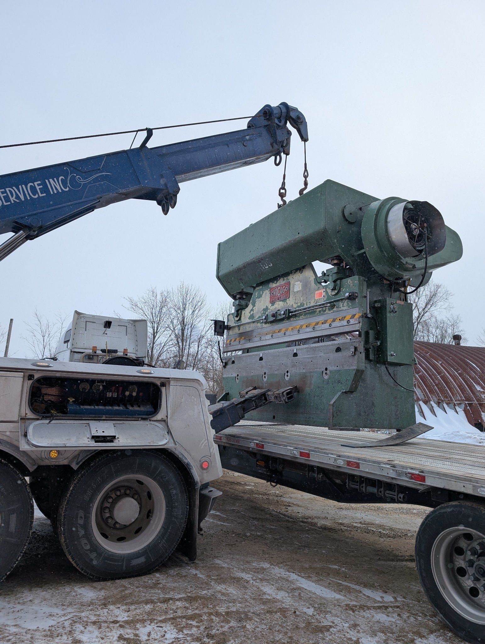 Crane lifting a large, green industrial machine onto a flatbed trailer in a snowy, outdoor setting.
