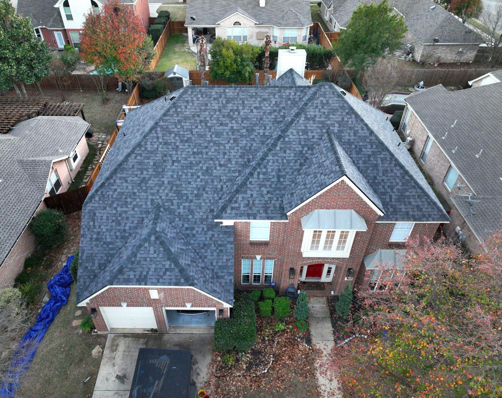 Aerial view of a brick house with a dark gray roof in a suburban neighborhood with fall foliage.