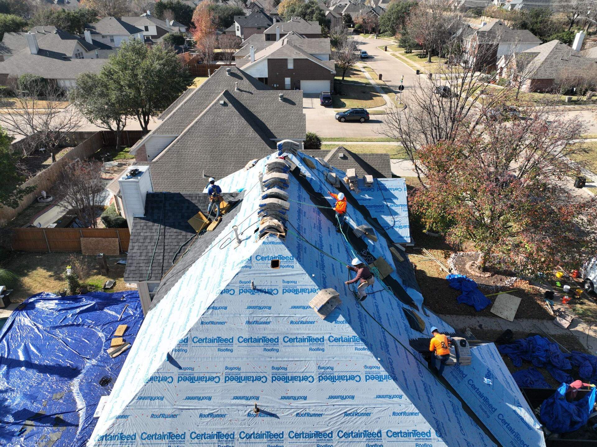 Roofers on a residential roof installing shingles, blue tarp covering part of the roof, sunny day.