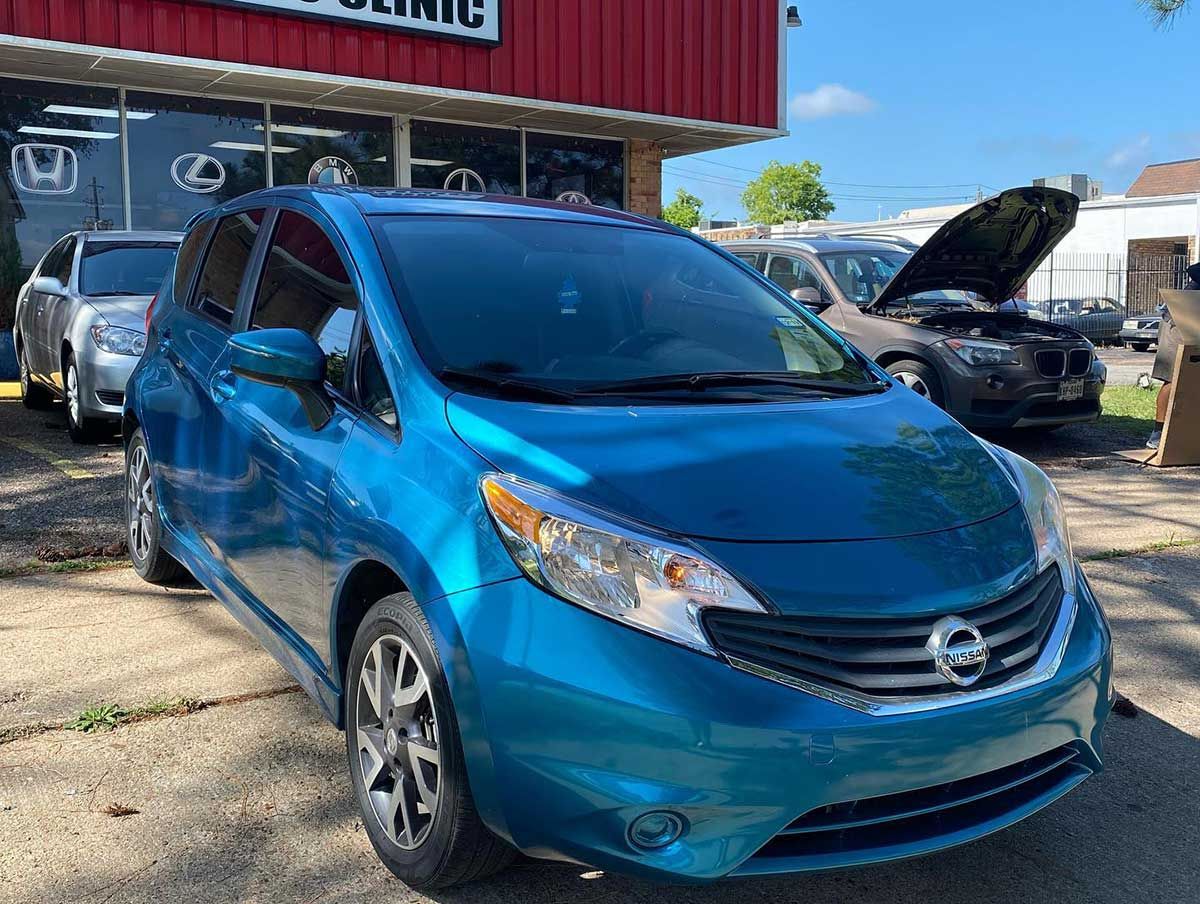 A blue nissan note with its hood up is parked in front of a car dealership.