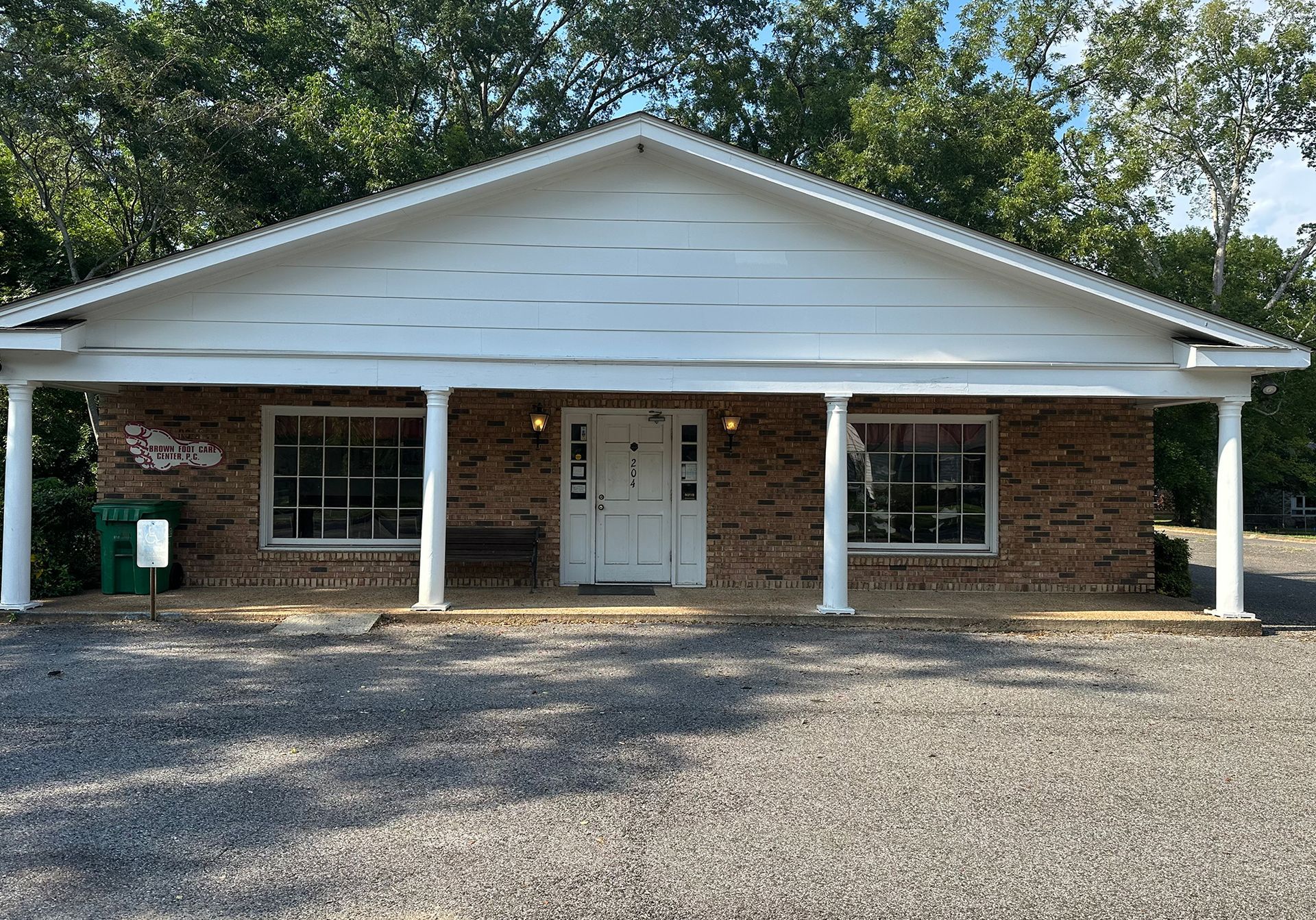 Brick building with white trim, columns, and a pitched roof. Gravel driveway, trees in the background.