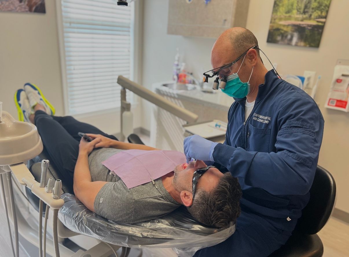 A dentist wearing a mask, gloves, and glasses, examining a patient in a dental chair.