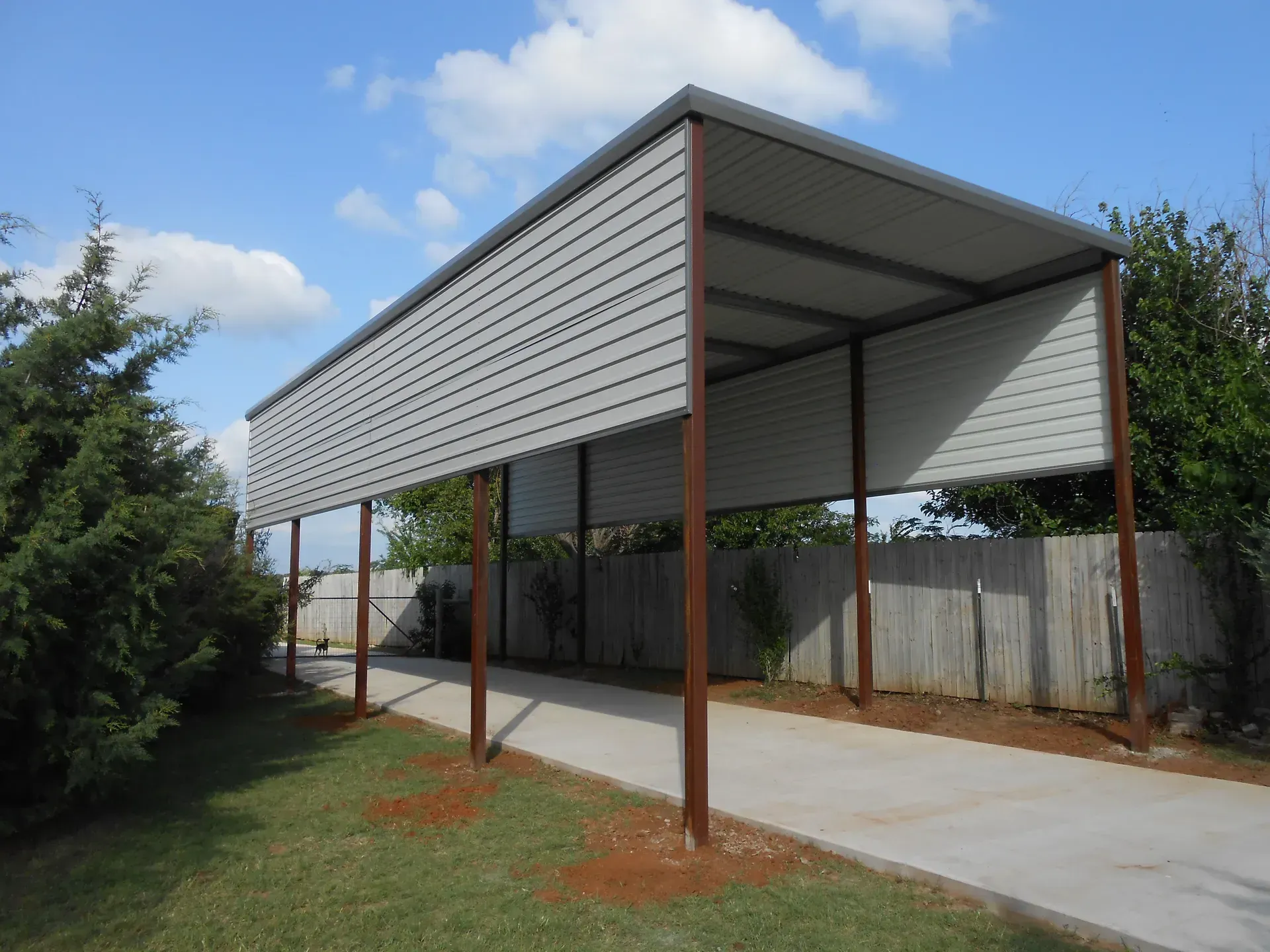 Covered driveway with a metal roof and support posts beside a fence and grassy yard