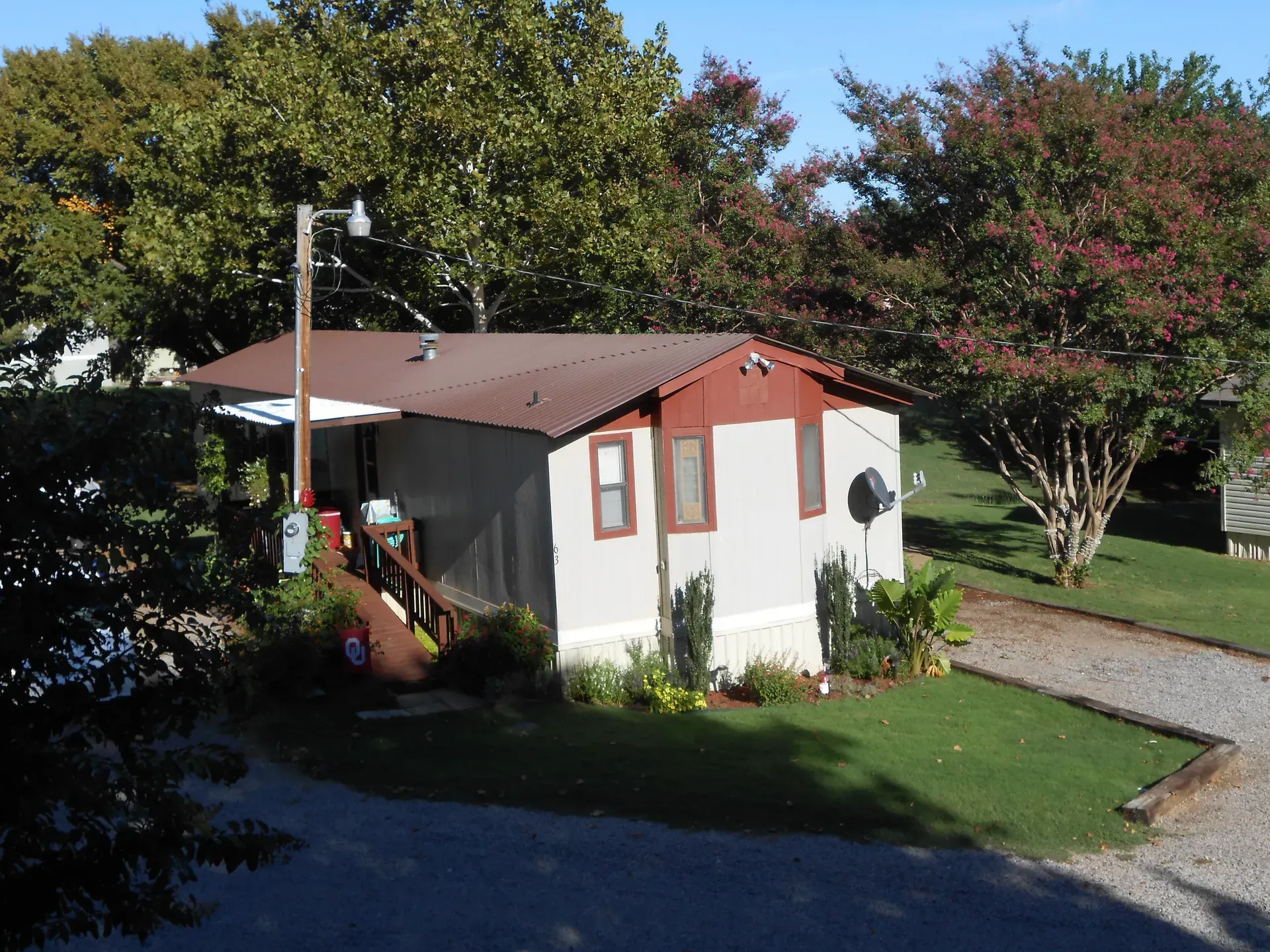 Small white shed with brown roof in a sunny garden, surrounded by trees and a gravel path.