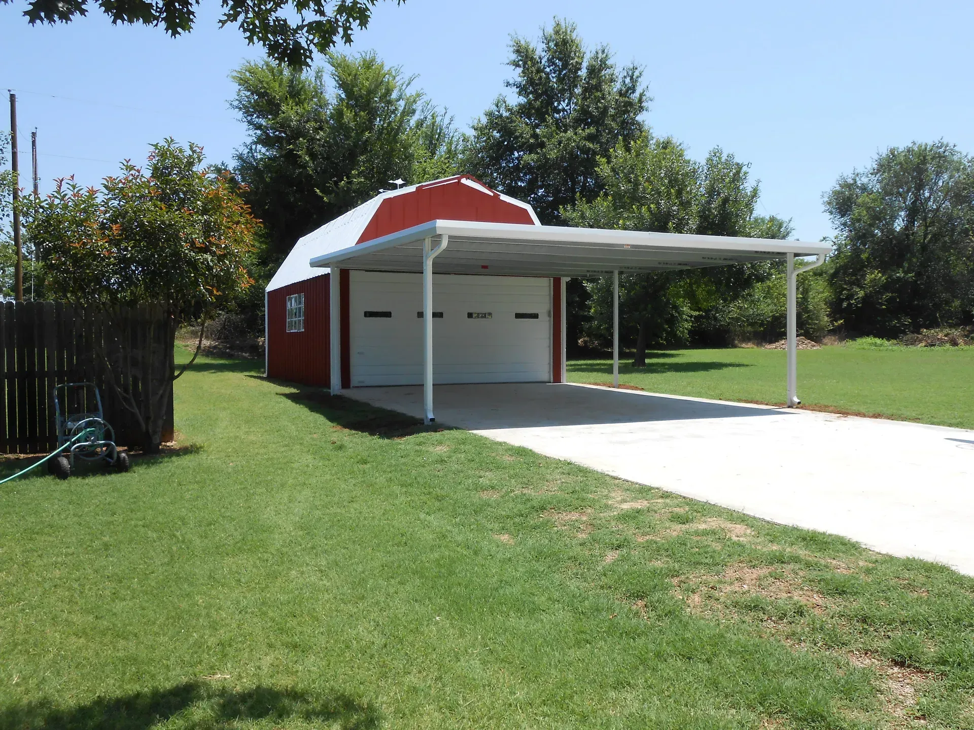 Red barn-style garage with white doors and carport beside a concrete driveway on a grassy lawn.