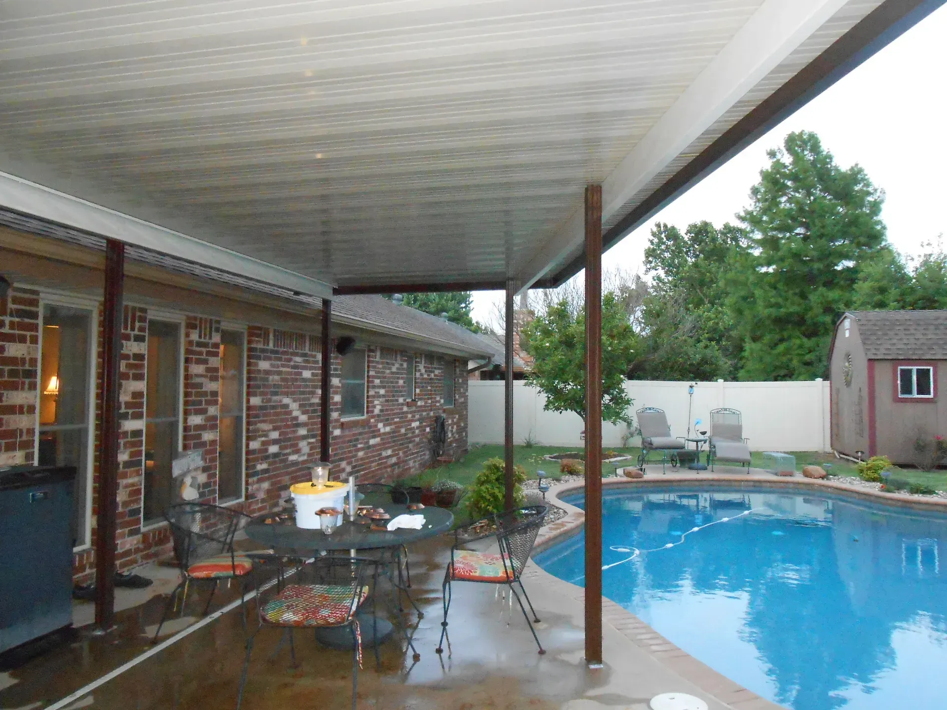 Covered patio beside a backyard pool with table, chairs, and brick house in the background