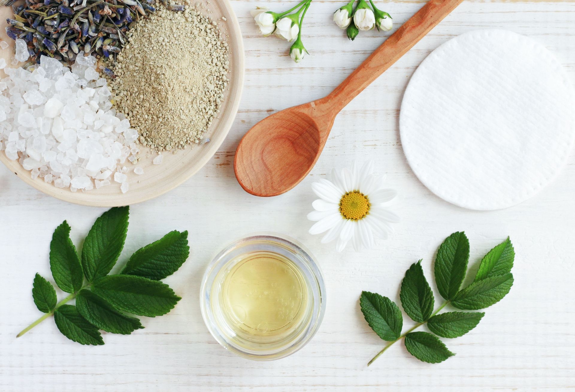 Spa treatment ingredients: salt, herbs, oils, flowers, wooden spoon, cotton pads, green leaves on white.