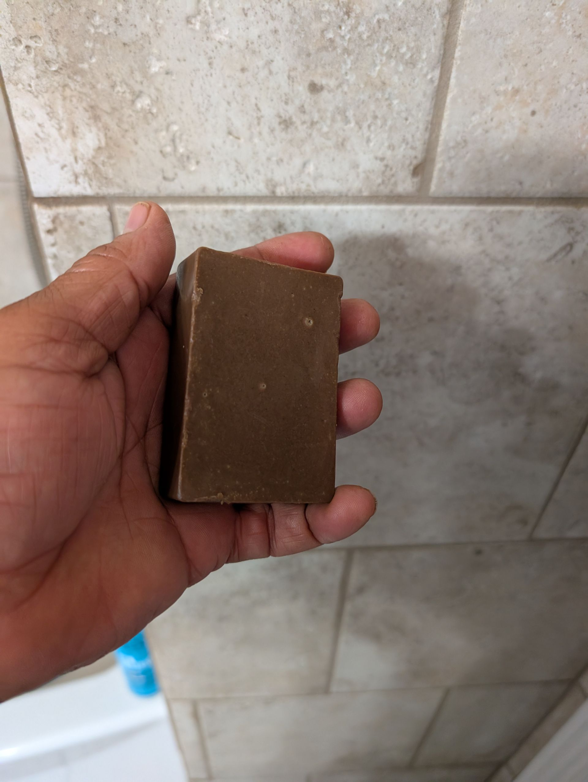 A hand holding a rectangular block of brown, textured soap in front of a tiled bathroom wall.