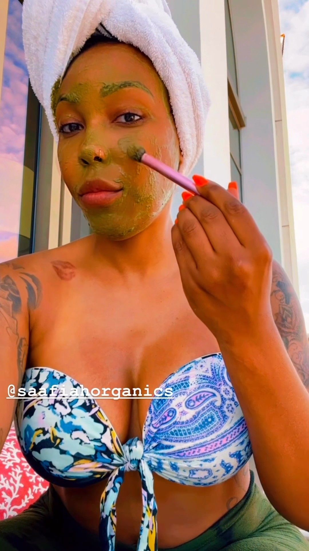 Woman applying a green face mask outdoors, wearing a bikini top and towel, holding a brush.