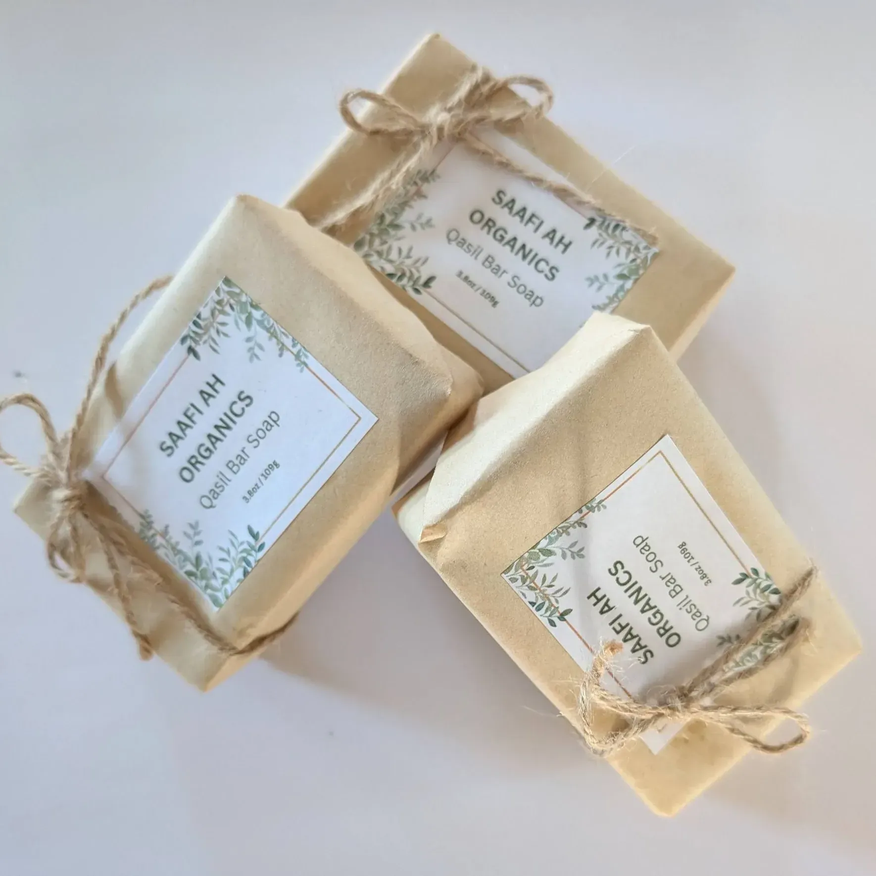 Three bars of Saafiah Organics Qasil soap wrapped in brown paper, tied with twine, and arranged on a white background.