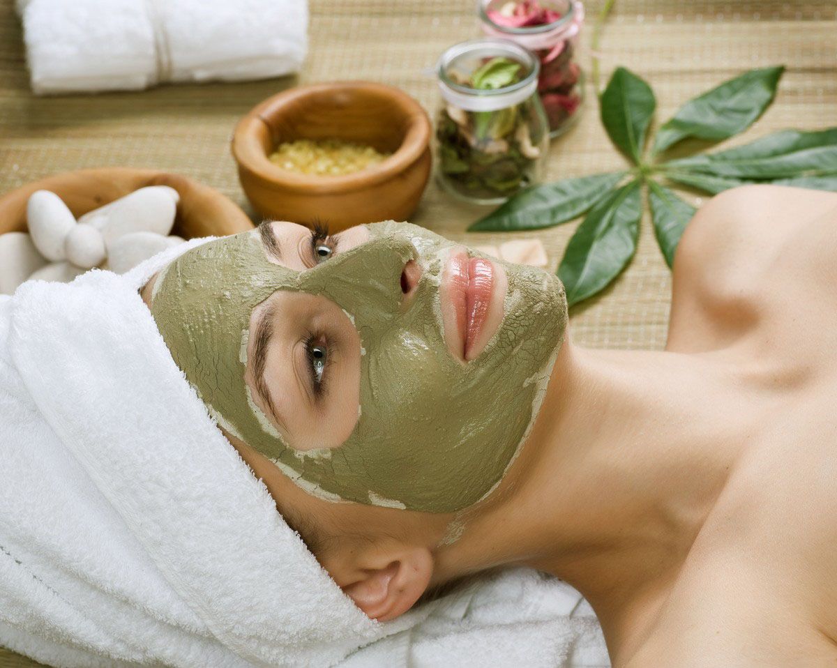 Woman with a green face mask, white towel on head, relaxing at a spa.