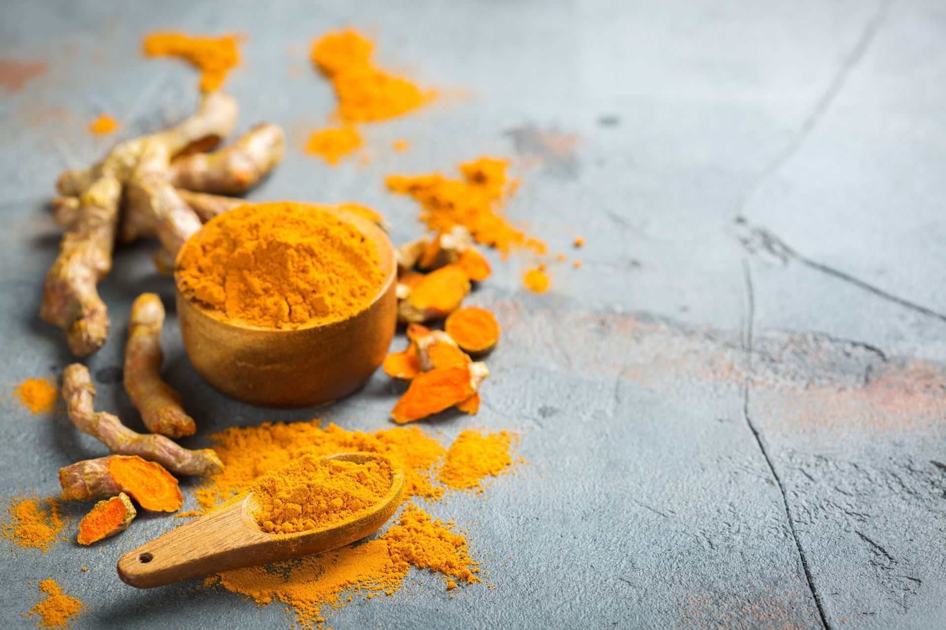 Turmeric powder in a wooden bowl and on a spoon, with fresh turmeric root pieces on a gray surface.